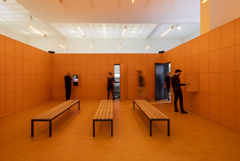 People move around an orange room containing three orange benches in the center.